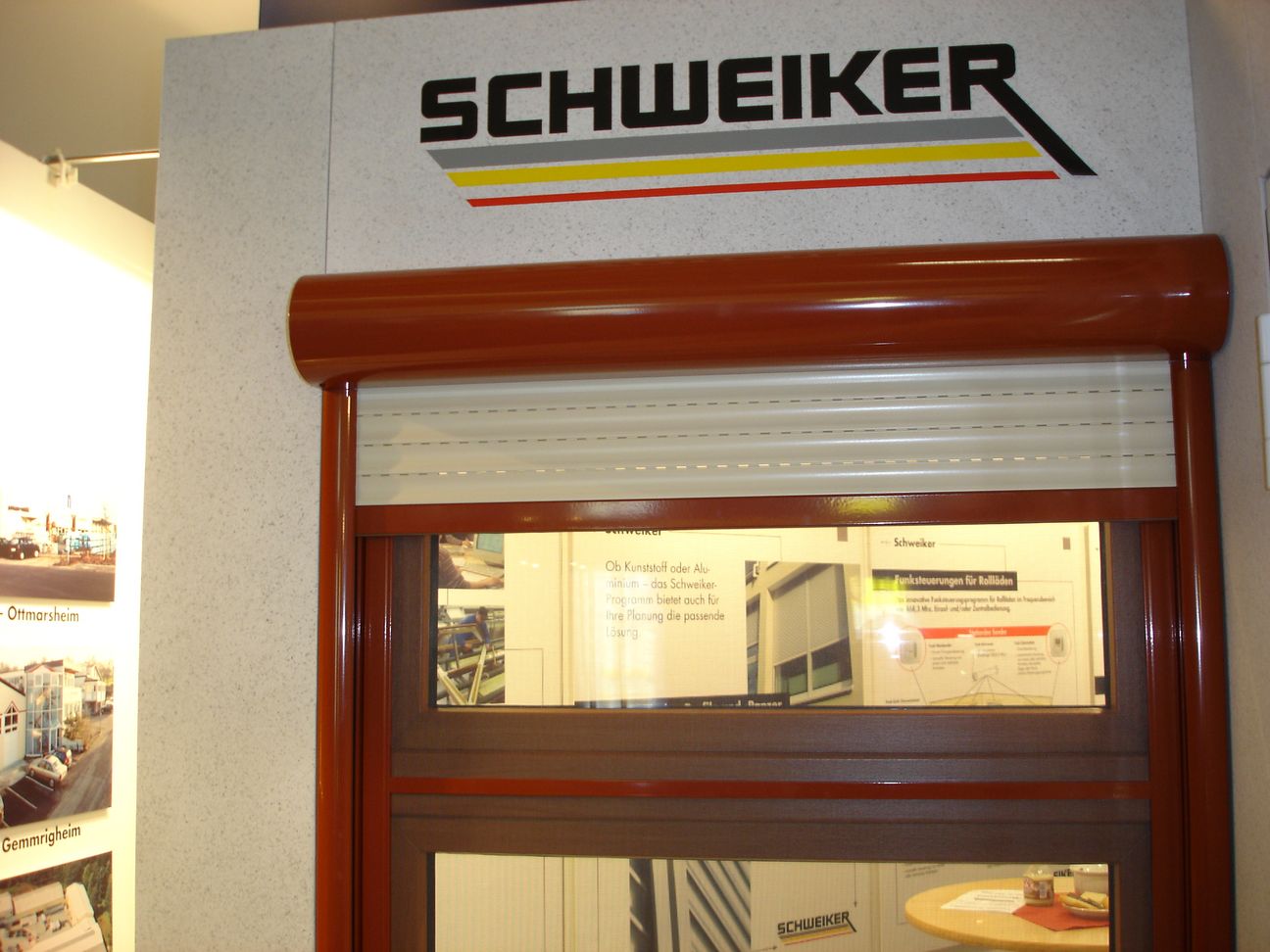 Ekspozycja okienna z częściowo opuszczoną, jasną osłoną przeciwsłoneczną, brązowa rama, logo producenta na górze, plakaty informacyjne widoczne w tle.